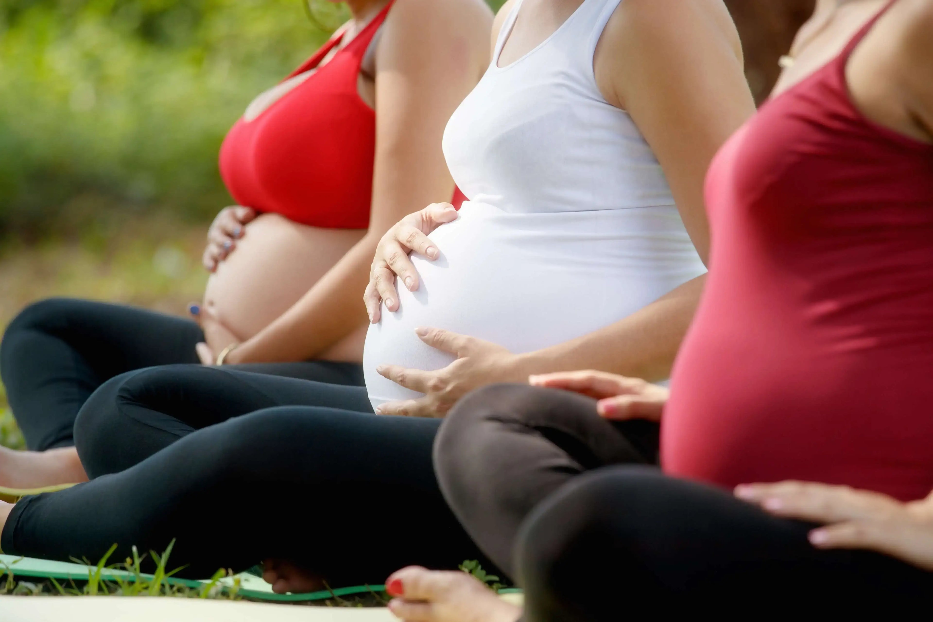 Three pregnant women sit together, the closest is wearing a red tank top and black pants, the middle is wearing a white tank top and holding her stomach, and the farthest is wearing a red bra holding her stomach with a green background