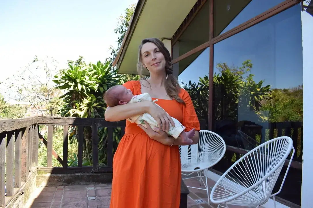 A blonde woman in an orange dress holds a newborn baby in a light jumper on an outdoor patio with white chairs and greenery in the background