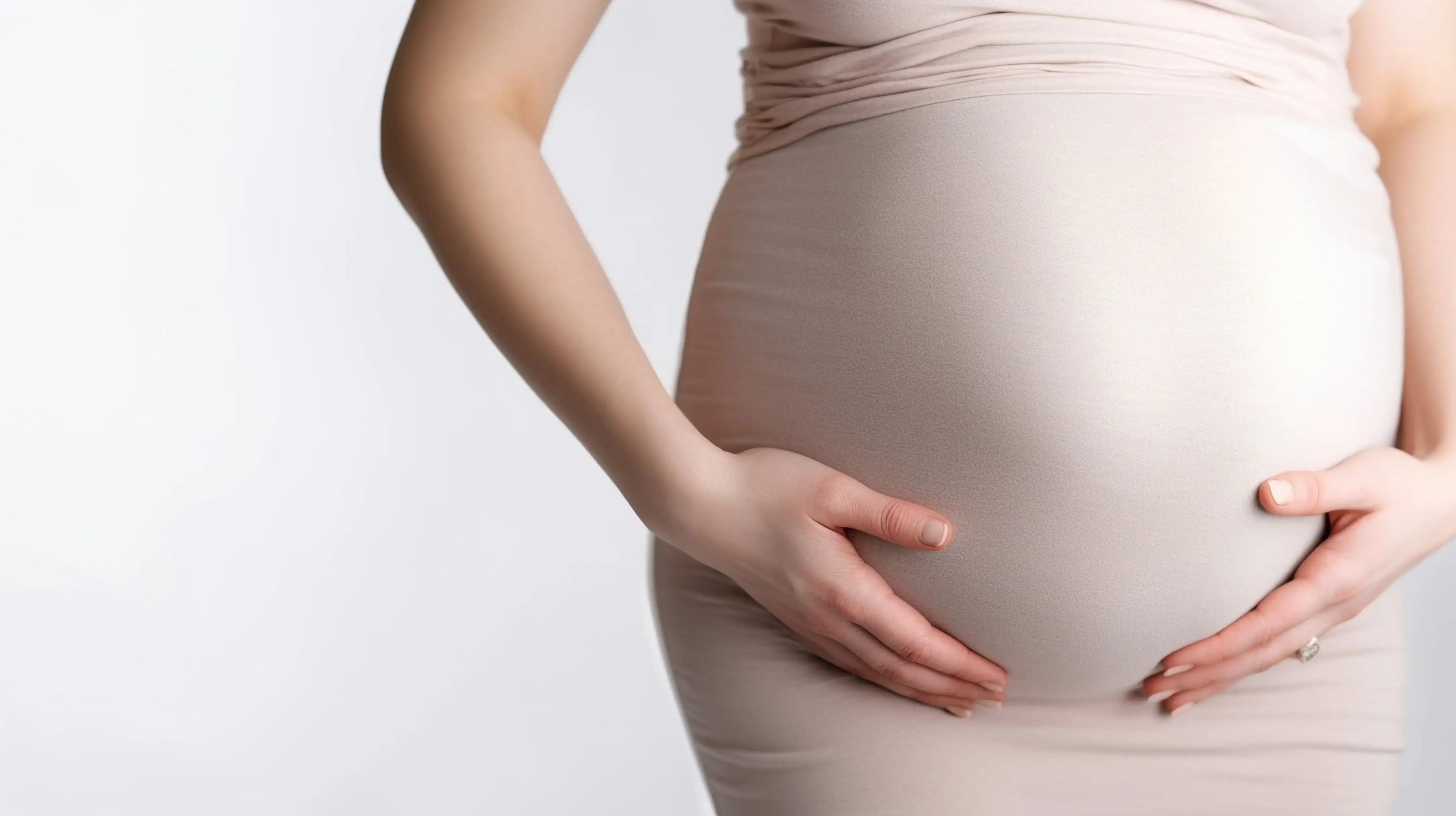 A pregnant woman wearing a light pink dress holds her pregnant stomach with both hands on a white background