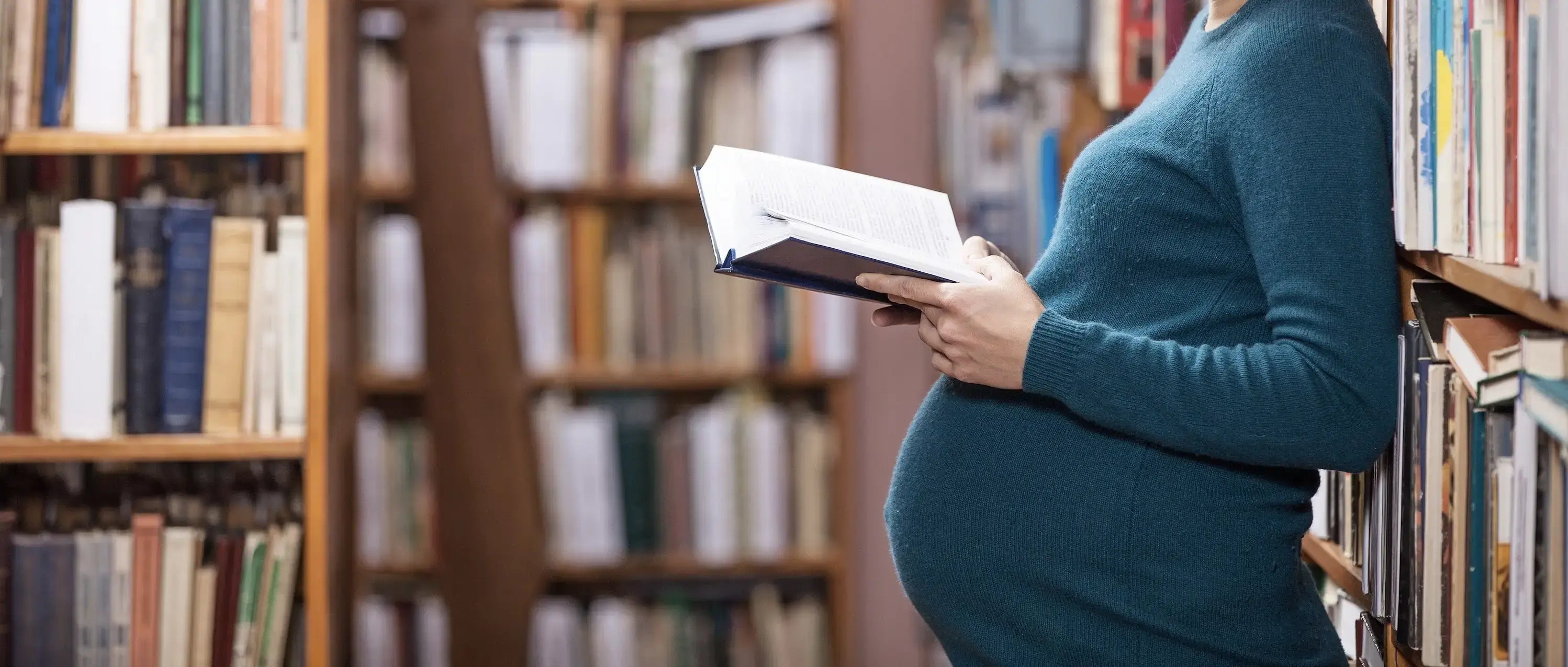 A pregnant woman dressed in green holds an open book in a library