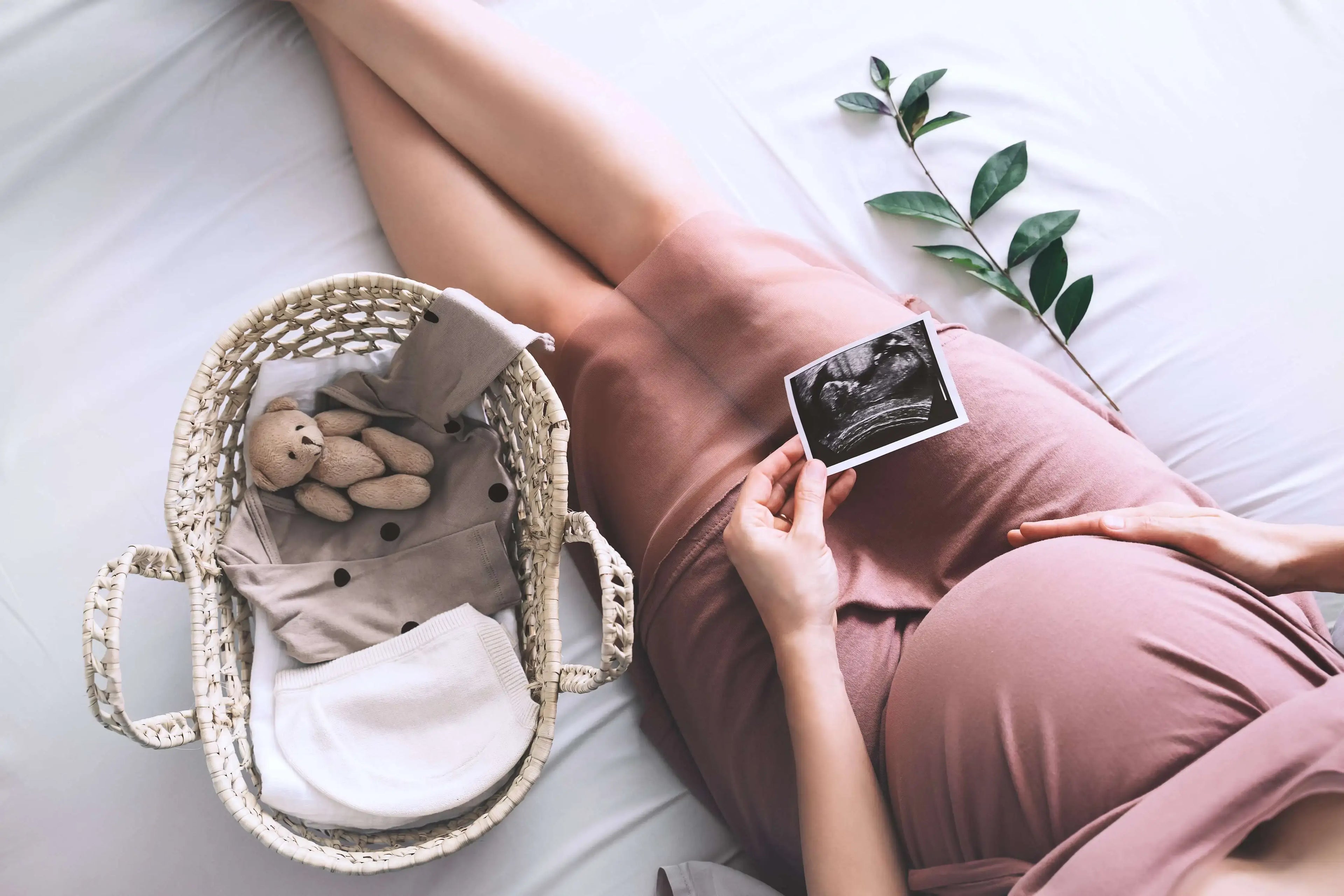 A pregnant woman in a mauve dress sits on a white bed with her hand on her stomach holding an ultrasound of her baby with greenery and a woven basket holding baby items nearby