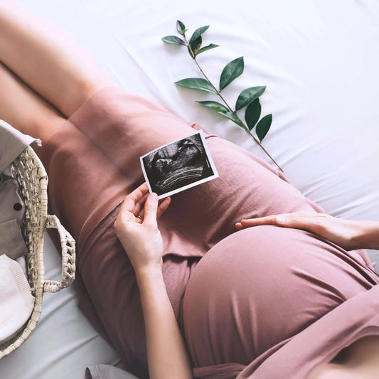 Pregnant woman in a mauve dress sits on a white bed touching her stomach and holding an ultrasound of her baby with a woven basket and greenery nearby