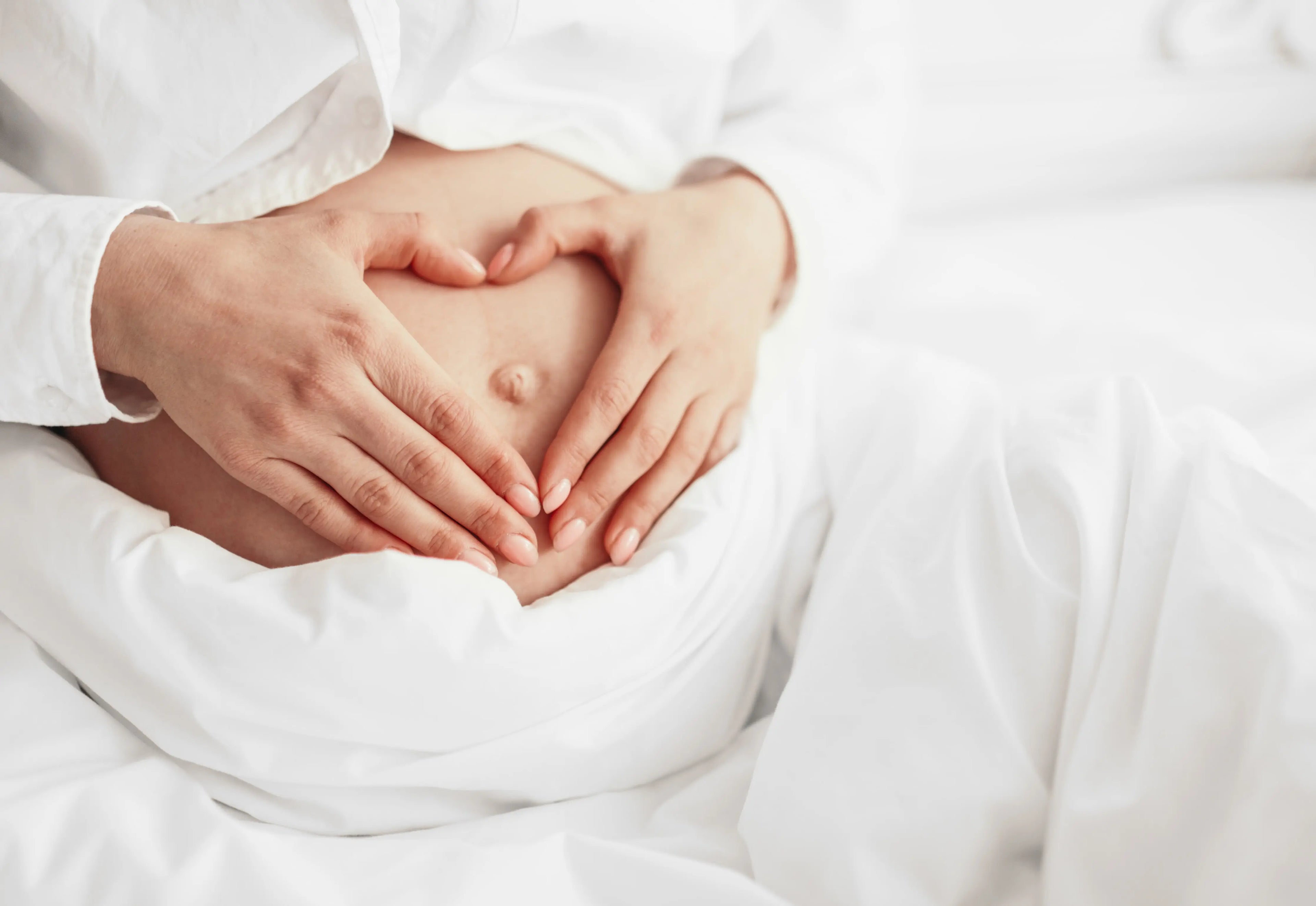 A pregnant person dressed in white holds their stomach with both hands forming a heart shape over the bellybutton 