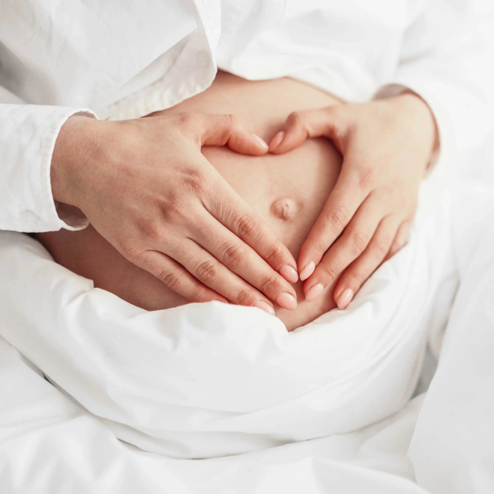 A pregnant person dressed in white holds their stomach with both hands forming a heart shape over the bellybutton