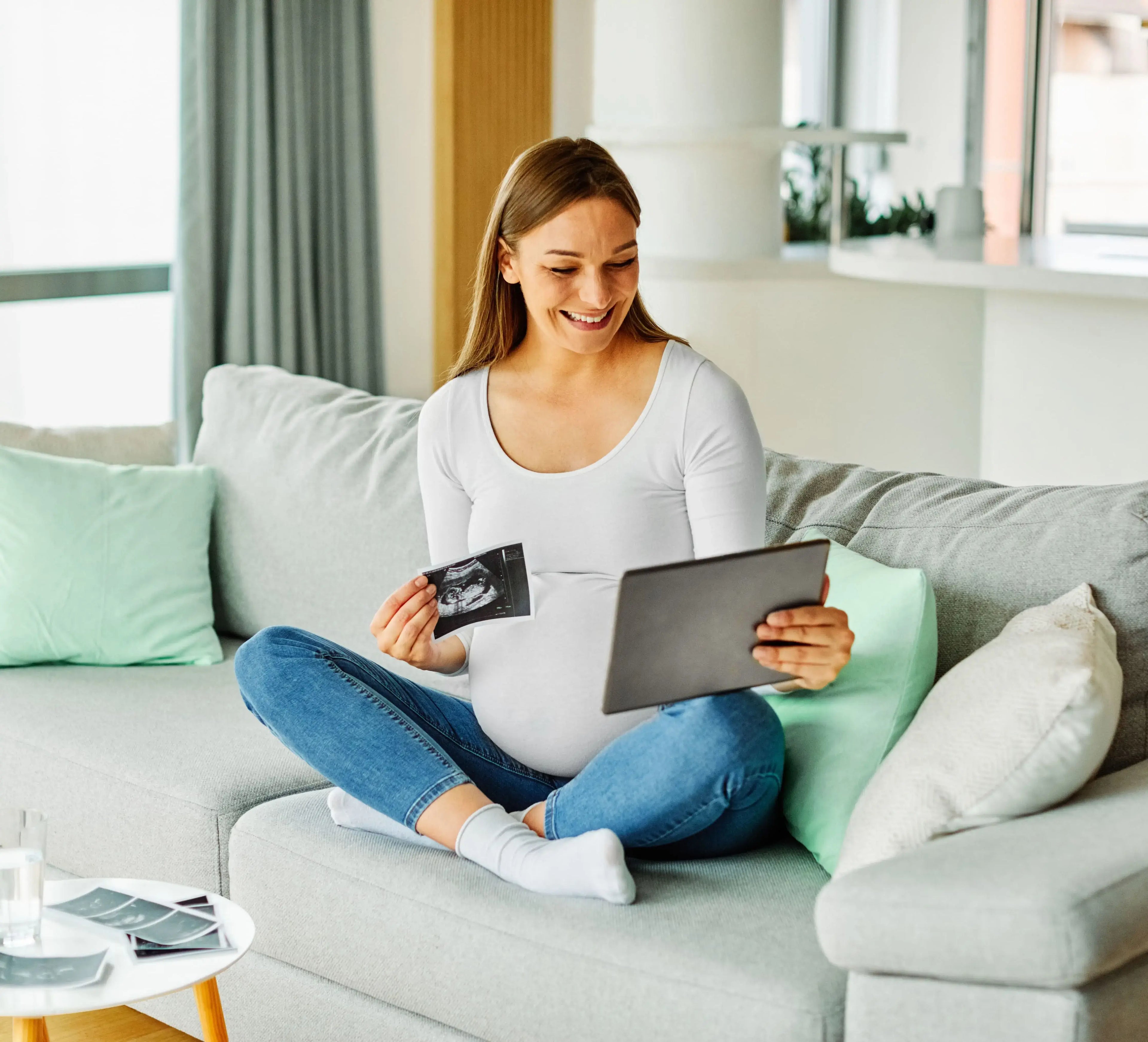 A blonde pregnant woman sitting on a light green couch smiles at her tablet holding an ultrasound with more on a nearby table