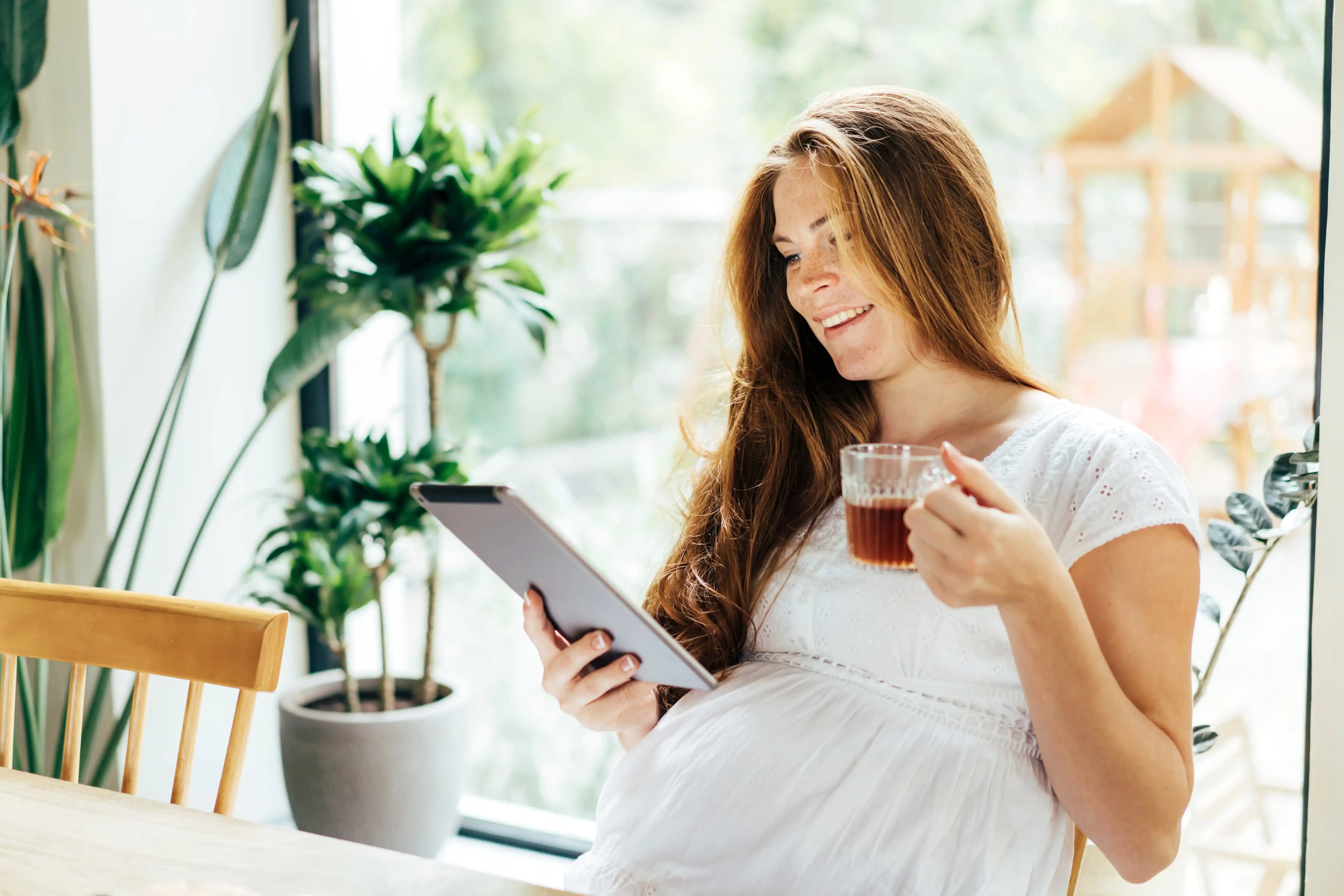 A brunette pregnant woman drinks from a teacup and smiles at a tablet while sitting at a table in a sunny room with plants in the background