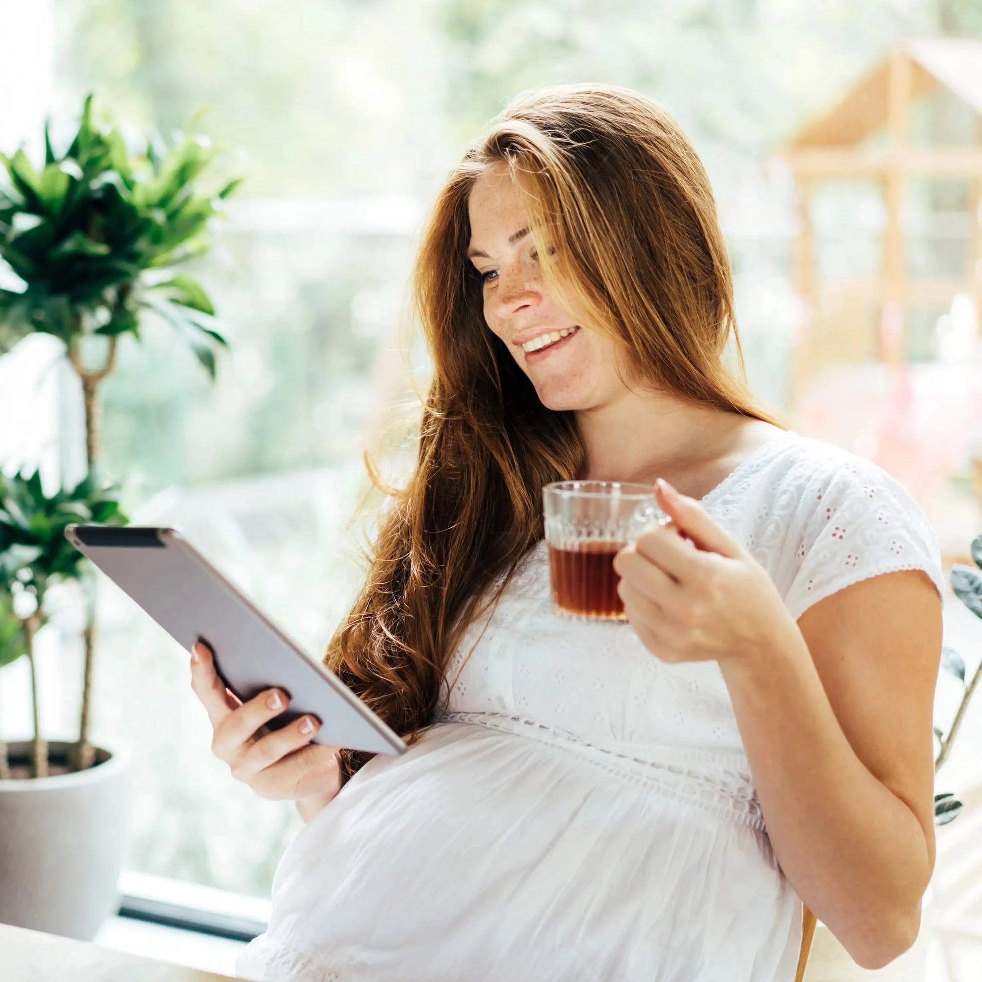 A brunette pregnant woman drinks from a teacup and smiles at a tablet while sitting at a table in a sunny room with plants in the background